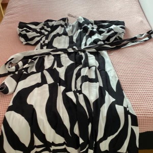 Svartvit omlottklänning från H&M - Säljer en svartvit omlottklänning från H&M i storlek XXL. Klänningen har ett coolt zebramönster, korta puffärmar och medföljande bälte i midjan. Materialet känns mjukt och lätt, perfekt för dig som gillar statement-plagg med unik design.