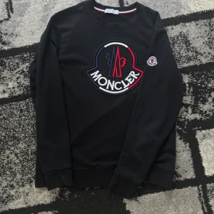 Svart moncler tröja - Svart sweatshirt från Moncler med stor broderad logga i vitt, rött och blått på bröstet samt mindre logga på ärmen. Klassisk rund halsringning och ribbade muddar. Perfekt för dig som gillar streetwear och vill sticka ut med en ikonisk look. Storleken är M, men ja skulle säga S.