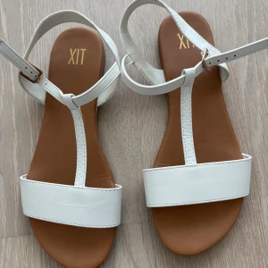 Vita sandaler från XIT - Snygga vita sandaler från XIT med bred rem över tårna och smalare rem med spänne runt ankeln. Sulan är brun och remmarna har en lätt textur. Perfekta för varma dagar och enkel att matcha med sommarens outfits.