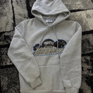 Trapstar hoodie - Snygg grå hoodie från Trapstar med broderad London Shooters-logga och pantermotiv på bröstet. Tröjan har huva med snörning och en klassisk känguruficka framtill. Perfekt för dig som gillar streetwear och vill sticka ut med coola detaljer.