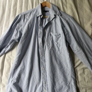 Randig skjorta från Mille Notti M - Snygg skjorta från Mille Notti i storlek M. Skjortan är vit med tunna blå ränder, har klassisk krage, bröstficka och knäppning framtill. Tillverkad i mjuk bomull, perfekt för en clean och stilren look.