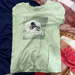 Ljusgrön t-shirt med California-tryck - Snygg ljusgrön t-shirt med coolt tryck av en cyklist och texten 'California State' på bröstet. T-shirten har klassisk passform och är kortärmad, perfekt för dig som gillar en avslappnad stil. Materialet känns mjukt och skönt mot huden.