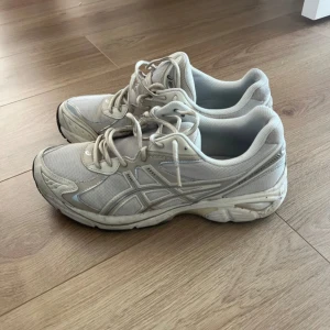 Vita sneakers från Asics - Säljer ett par fräscha vita Asics sneakers med mesh och syntetdetaljer. Skorna har snörning och en chunky sula med svarta och vita partier undertill. Perfekta för dig som gillar sportig stil och vill ha bekväma skor till vardagen.