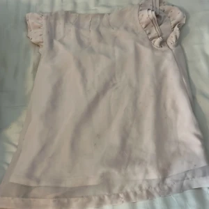 Ljusrosa blus med volangärmar - Supersöt ljusrosa blus med volangdetaljer på ärmarna och knappar framtill. Blusen har en lös passform och är ärmlös, perfekt för varmare dagar. Tillverkad i ett tunt och mjukt material som känns skönt mot huden. 