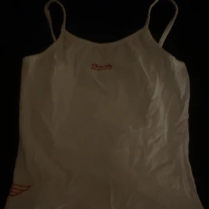 Vit linne-topp från Trespass med hjärtdetalj - Vit linne-topp från Trespass med tunna axelband och röd logga framtill. Nedtill finns en cool röd hjärtdetalj med vingar. Toppen är gjord i mjuk bomull med lite stretch för extra komfort. Perfekt för en chill sommarlook eller lager-på-lager.