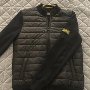 Svart bomberjacka från Barbour M - Svart bomberjacka från Barbour International med quiltad framsida och slät baksida. Jackan har ribbad krage med svarta och gula ränder, dragkedja framtill och Barbour-patch på ärmen. Snygg, sportig look med två sidofickor och långärmad design.