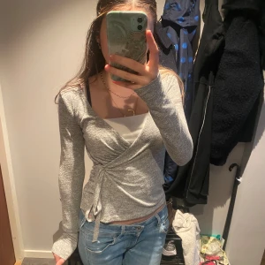 Grå omlottopp med knyt - Säljer en grå omlottopp med långa ärmar och knyt i sidan. Toppen har en v-ringad design och är i mjukt, stretchigt material. Perfekt att styla med jeans eller kjol för en chill look.