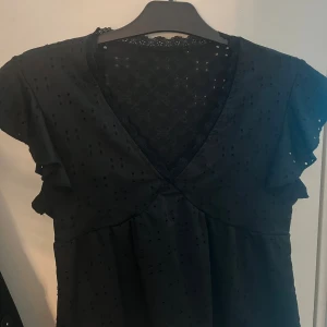 Svart kortärmad klänning med spets - Svart klänning med v-ringning och fina spetsdetaljer upptill. Kortärmad modell med volang vid ärmarna och diskret broderat mönster i tyget. Perfekt för dig som gillar en romantisk och stilren look.