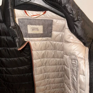 Svart pufferjacka från Camel Active - Snygg svart pufferjacka från Camel Active med quiltad design och hög krage. Jackan har dragkedja framtill, två stora fickor med knappar och en bröstficka med dragkedja. Insidan är ljusbeige och materialet är syntet, perfekt för kyliga dagar.