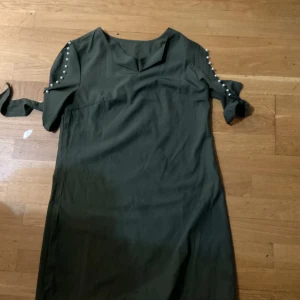 Mörkgrön klänning med pärldetaljer - Snygg mörkgrön klänning med pärldetaljer längs ärmarna och knytband vid ärmsluten. Klänningen har en rak passform och kort ärm, perfekt för dig som gillar stilrena plagg med en twist. Minns inte vart den är köp men jätte söt på och bekväm 