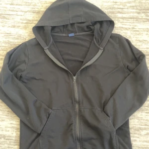 Svart hoodie med dragkedja - En klassisk svart hoodie med dragkedja och huva. Tröjan har långa ärmar, känguruficka framtill och är tillverkad i mjuk bomull. Perfekt för chill dagar eller när du vill ha en enkel och stilren look.