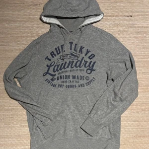 Grå hoodie från Tokyo Laundry - Säljer en grå hoodie från Tokyo Laundry med blå tryck framtill. Tröjan har huva med snörning och en stor magficka. Perfekt för dig som gillar en avslappnad och sportig stil. Mjuk bomullsblandning och långa ärmar. Kontakta vid frågor!