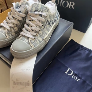 Dior B23 sneakers med monogram - Snygga Dior B23 sneakers i vitt och grått med ikoniskt Dior-monogram över hela skon. Lågt skaft, vita snören och robust vit sula med grå detaljer. Ovandel i syntetmaterial och textil, med tydlig Dior-logga på sidan. Kommer med originalkartong och dustbag.