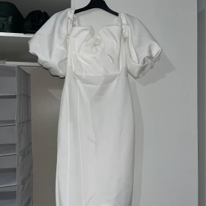 Vit kortklänning med puffärmar - Supersnygg vit kortklänning med stora puffärmar och unik draperad framsida. Klänningen har en modern och trendig look med fyrkantig urringning och är tillverkad i ett slätt, satinliknande material. Perfekt för dig som vill sticka ut med en cool och elegant stil.
