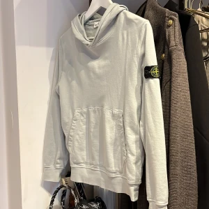 Ljusgrå hoodie från Stone Island - Snygg ljusgrå hoodie från Stone Island med klassisk patch på vänster ärm. Tröjan har huva, magficka och långa ärmar. Tillverkad i mjuk bomull för en skön känsla. Perfekt för dig som gillar streetwear och vill ha en stilren look. Kvitto finns den är köpt på NK och är använd ungefär 1-2 gånger 