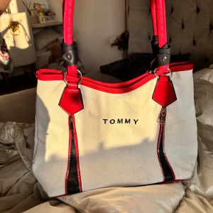 Vit och röd axelväska från Tommy - Stor och rymlig axelväska från Tommy i vit canvas med röda och svarta detaljer. Perfekt för universitet eller gymnasiet eftersom det rymmer mycket. Väskan har två långa röda handtag, dragkedjor framtill och ett stort huvudfack med innerficka. Snygg kontrast mellan färgerna och coolt broderad logga på framsidan. Lite smutsig 