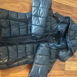 Svart pufferjacka från Lager157, XS - Säljer en blå pufferjacka från Lager157 i storlek XS. Jackan har huva, dragkedja framtill och två  fickor med dragkedja. Glansigt tyg. Jättevarm och skön. 