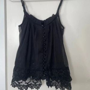 Svart genomskinlig spetslinne från Vila XS - Supersött svart linne från Vila med tunna axelband, dekorativa knappar framtill och bred spetskant nertill. Luftigt tyg med lätt struktur och romantisk känsla. Perfekt att styla med jeans eller kjol för en cool look.