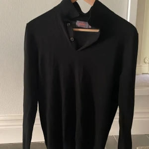 Svart knappad merinoullströja från Morris - Snygg svart långärmad tröja från Morris i 100% merinoull. Tröjan har hög krage med två knappar och stilren design som passar perfekt till ett par jeans. Skön och mjuk känsla, perfekt för kall sommarkväll.