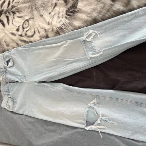 Ljusblå ripped jeans från Gina Tricot - Säljer ett par ljusblå jeans från Gina Tricot med raka ben och stora slitningar på båda knäna. Klassisk femficksmodell med dragkedja och knapp. Perfekta för en avslappnad och trendig look.