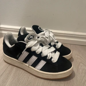 Adidas Campus 00’s sneakers svart/vit 38 - Svarta Adidas Campus sneakers med klassiska vita ränder och breda vita skosnören. Ovandel i mocka och sula i gummi. Endast använt 2-3 ggr. Säljes pga för liten storlek. Pris går självklart att diskuteras☺️