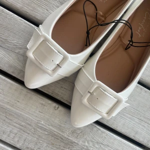 Vita ballerinaskor med stor spännen🤍 - Snygga vita ballerinaskor från New Look med spetsig tå och bred passform. Skorna har en stor dekorativ spänne framtill och är gjorda i glansigt syntetmaterial. Säljer med prislappen på - skulle igentligen använda till studenten men var för stora. Perfekt för dig som har storlekar 39 skulle jag säga. De står att de är en bred passform, men skulle gissa att dem passar en vanlig 39 (om du inte har superduper smala fötter såklart haha😇