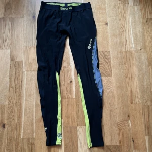 Svarta träningstights från Skins - Snygga svarta träningstights från Skins med limegröna och grå detaljer längs benen. Tightsen har en elastisk midja med logga och är perfekta för dig som gillar att träna. Materialet är stretchigt och sitter tight mot kroppen. Endast provade men har för låg midja för min smak. Nypris kring 1000kr