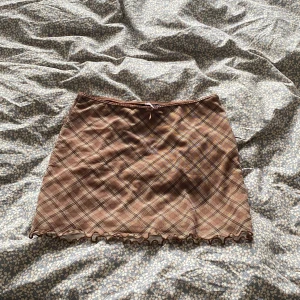 Brun rutig kortkjol med volangkant - Supersöt brun rutig kortkjol med volangkant längst ner och liten rosett i midjan. Kjolen har en slim passform och är perfekt för dig som gillar en trendig och lekfull stil.