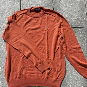 Rostbrun merinoullströja från Morris - Snygg rostbrun stickad tröja från Morris i 100% merinoull. Klassisk rund halsringning och diskret broderad logga på bröstet. Perfekt för dig som gillar stilrena och bekväma plagg med en modern touch.