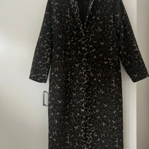 Leopardmönstrad kappa från NA-KD - Snygg lång kappa från NA-KD i leopardmönster med svarta och bruna toner. Klassisk krage, knäppning med två knappar framtill och raka ärmar. Perfekt statement-plagg för dig som vill sticka ut i höst och vinter.
