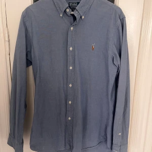 Blå skjorta Polo Ralph Lauren slim fit - Säljer en stilren blå skjorta från Polo Ralph Lauren i slim fit-modell. Skjortan har klassisk button down-krage, broderad logga på bröstet och vita knappar. Tillverkad i mjuk bomull och har långa ärmar. Perfekt för dig som gillar en clean och tidlös look.
