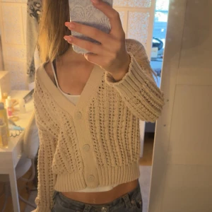 Beige stickad kofta med knappar - Säljer en beige stickad kofta med v-ringning och stora knappar framtill. Koftan är croppad och har ett luftigt, hålmönstrat stickat material. Perfekt att slänga över ett linne eller topp för en chill look.