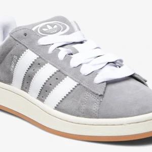Adidas Campus grå sneakers mocka - Adidas Campus sneakers i grå mocka med klassiska vita ränder på sidorna och vit logga på plösen. Skorna har vit sula med gummidetalj och breda vita skosnören. Perfekt för dig som gillar retrostil och streetwear. INTE ÄKTA!!