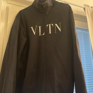 Svart vindjacka från Valentino VLTN - Svart tröja från Valentino med VLTN-tryck i vitt på bröstet. Tröjan har hög krage, hel dragkedja framtill och långa ärmar. Storlek är Medium i herrstorlek.