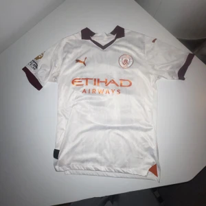 Manchester City De Bruyne tröja Puma - Säljer en vit Manchester City fotbollströja från Puma med De Bruyne och nummer 17 på ryggen. Tröjan har korta ärmar, vinröda detaljer vid krage och ärmslut, samt orange Etihad Airways-tryck på bröstet. Materialet är lätt och andas, perfekt för match eller träning.