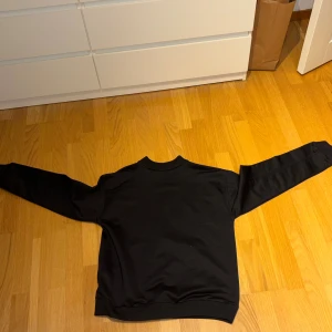 Svart ziptröja från LMTD, storlek 176 - Svart långärmad ziptröja från LMTD med stilren design och ribbad krage. Tröjan har en hel dragkedja framtill och två fickor. Materialet är mjukt och känns som bomull, perfekt för chill dagar. Passar dig som gillar enkel och clean stil. Använd kanske 3 gånger. 