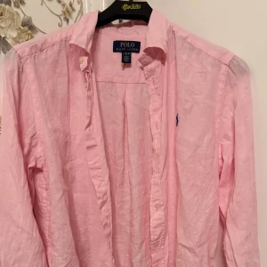 Rosa linneskjorta från Polo Ralph Lauren - Snygg rosa skjorta från Polo Ralph Lauren i storlek L. Tillverkad i lätt och luftig linne, perfekt för varmare dagar. Klassisk krage, långa ärmar och knappar framtill. Ikoniska blå Polo-loggan broderad på bröstet ger en stilren touch.