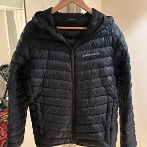 Blå dunjacka Peak Performance M - Blå dunjacka från Peak Performance i storlek M. Jackan har huva, dragkedja framtill och diskret logga på bröstet och ärmen. Den är quiltad med horisontella sömmar och har en cleani, sportig look som funkar till de flesta stilar. Jackan är i super gott skick och har inte blivit använd särskilt mycket. 