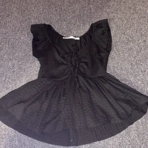 Svart blus med knyt från 157 XS - Säljer en svart blus från 157 i storlek XS. Blusen har korta ärmar med volang, rund halsringning och snygg knytning framtill. Materialet är lätt och har en subtil struktur som ger en cool vibe. Perfekt för dig som gillar en feminin och trendig look. Aldrig använd 