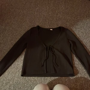 Svart ribbad omlottopp från H&M Divided - Svart ribbad omlottopp från H&M Divided i storlek S. Toppen har lång ärm, v-ringning och snygg knytning framtill. Materialet är mjukt och stretchigt, perfekt för att skapa en chill och stilren look. Passar dig som gillar enkla detaljer och svart basic.