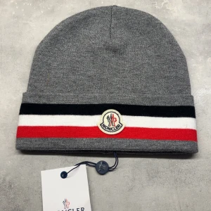 Grå Moncler mössa med ränder - Snygg grå mössa från Moncler med klassiska svarta, vita och röda ränder runt kanten. Mössan har Moncler-logga framtill och är tillverkad i mjukt stickat material. Perfekt accessoar för att lyfta din vinterstil.