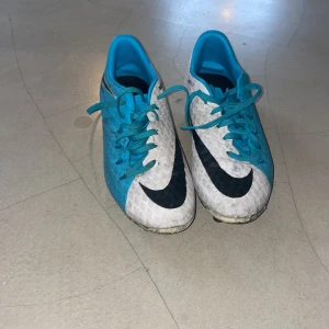 Nike blåvita fotbollsskor med dobbar som är väldigt bra skick och perfekta för dig som vill sticka ut på planen - Säljer ett par Nike fotbollsskor i blått och vitt med svarta detaljer. Skorna har snörning och rund tå, samt platt sula med fasta dobbar för gräsplan. Ovandelen är tillverkad i syntetmaterial med ett mönstrat yttre och stor Nike-logga på sidan. Perfekt för dig som vill sticka ut på planen.