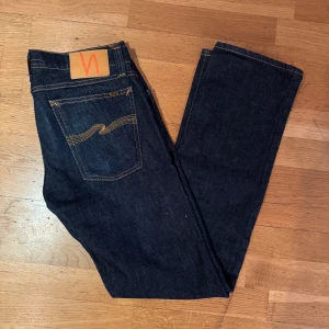 Mörkblå jeans från Nudie Jeans - Snygga mörkblå jeans från Nudie Jeans med klassisk femficksdesign och orangea sömmar. Modellen har rak/slim passform och normal midja, tillverkade i slitstarkt jeansmaterial. Bakfickorna har Nudies ikoniska broderade vågmönster och läderpatch med logga.