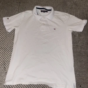 Vit pikétröja från Tommy Hilfiger - Klassisk vit pikétröja från Tommy Hilfiger med broderad logga på bröstet och ärmen. Tröjan har krage, knappar framtill och korta ärmar med ribbade muddar. Perfekt för en clean och stilren look.