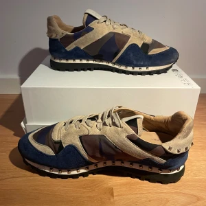 Valentino Rockrunner sneakers blå/beige - Snygga Valentino Rockrunner sneakers med beige mockadetaljer, blå och bruna partier i camouflage-mönster. Skorna har snörning, rund tå och platt sula med nitar längs skorna