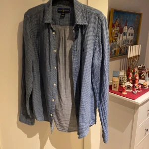 Blårutig skjorta från Ralph Lauren - Blårutig skjorta från Ralph Lauren med klassisk krage och knappar framtill. Skjortan har lång ärm och det ikoniska broderade Polo-logot på bröstet. Tillverkad i mjuk bomull som känns skön mot huden. Perfekt för dig som gillar stilren och tidlös design. En liten XL som passar perfekt på M