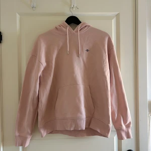 Ljusrosa hoodie från Gant - Säljer en ljusrosa hoodie från Gant med klassisk logga broderad på bröstet. Tröjan har huva med snörning, stor magficka och ribbade muddar vid ärmslut och nederkant. Perfekt för chill dagar och enkel att matcha med jeans eller mjukis.
