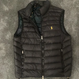 Svart dunväst från Polo Ralph Lauren - Svart dunväst från Polo Ralph Lauren med gul broderad logga på bröstet. Västen har quiltad design, två dragkedjeförsedda fickor och hög krage. Materialet är glansigt och känns lätt, perfekt för lager-på-lager. Stängs med dragkedja framtill. Ett litet hål på framsidan som är igenstängt med en lapp som knappt syns.
