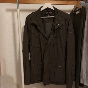 Mörkgrön Field jacka från Woolrich - Parkajacka från Woolrich i mörkgrön färg med flera fickor framtill, snygga knappar och dragkedja. Jackan har hög krage och detaljerade axelpartier. Materialet är slitstark polyester och jackan har lång ärm. Perfekt för dig som gillar funktionell och stilren design. Hör av dig vid frågor och priset är inte hugget i sten!