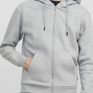 Grå hoodie  - Snygg ljusgrå hoodie från Jack & Jones med dragkedja framtill, huva med snörning och stora fickor. Perfekt för chill dagar eller när du vill ha en enkel och clean look. Tillverkad i mjuk bomullsmix för extra komfort.